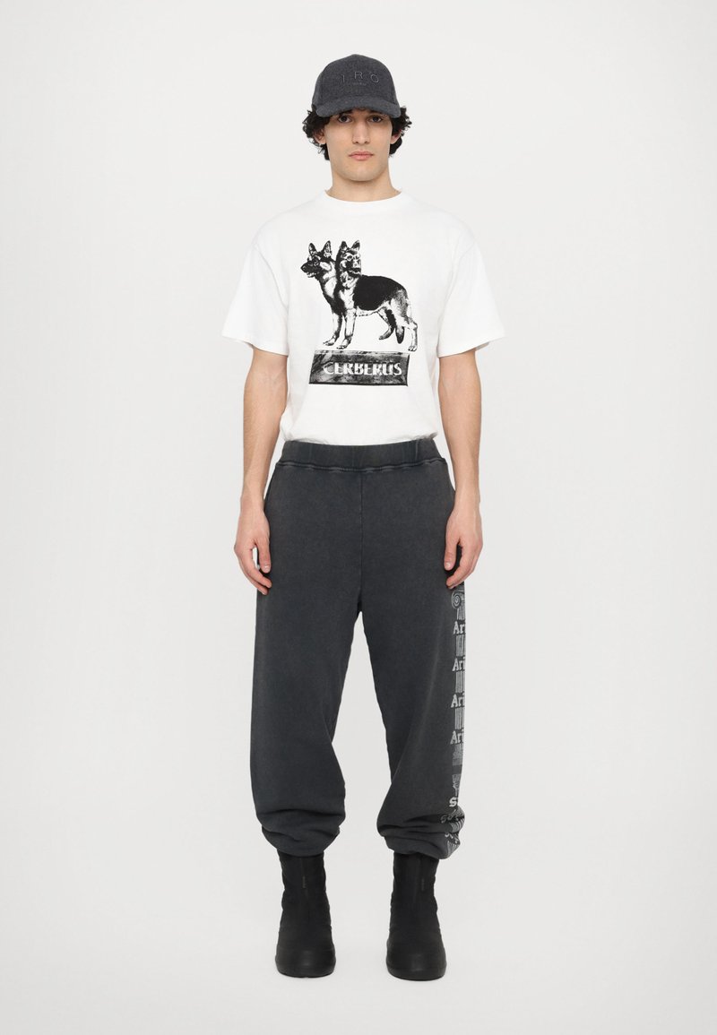 Weißes T-Shirt mit einem schwarzen Grafikdesign von Hunden, kombiniert mit dunkelgrauen Sweatpants, die seitlich mit Text bedruckt sind, und schwarzen Stiefeln.