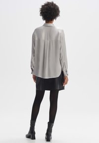 Chemise grise soyeuse à manches longues avec col, détail de pli au dos et ourlet arrondi, associée à une jupe en cuir noire et à des collants noirs.