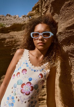 Maillot de bain blanc avec des imprimés colorés de coquillages et de fleurs, décolleté festonné, associé à des lunettes de soleil rectangulaires bleu clair et des cheveux bouclés.