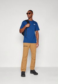 Modra kratka majica s kratkimi rokavi, bež hlače s potisnimi žepi z črnim zadrgo, črni nahrbtnik in črna obutev; sončna očala, nevtralna podlaga.