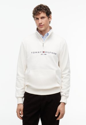 Junger Mann mit lockigem Haar, der einen weißen Tommy Hilfiger Quarter-Zip-Sweatshirt und dunkle Hose trägt, steht vor einem weißen Hintergrund.