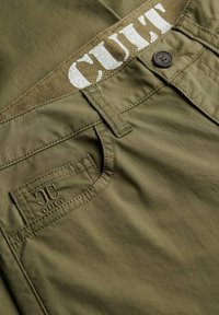 Close-up van olijfgroene broek met knoop, riemlussen en "JC CULT" geborduurd op het voorvak en de tailleband.