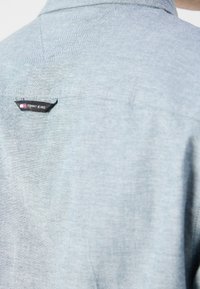 Vaaleansininen farkkutakki takaa, jossa on pieni musta "Tommy Jeans" -merkki ja näkyvät kauluksen sauman ompeleet.