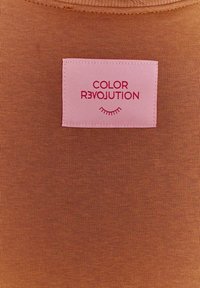 Hnědá ribbovaná látka zblízka s růžovou etiketou, na které je napsáno "COLOR R3VOLUTION" červeným písmem a malou grafikou řas.