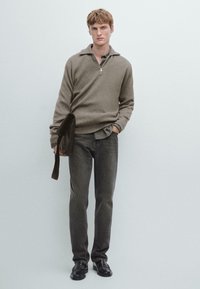 Beiger Reißverschluss-Pullover mit Strukturstrick, graue Jeans und schwarze Schuhe. Das Model hält eine dunkle Tasche, darunter ist ein kariertes Hemd sichtbar.
