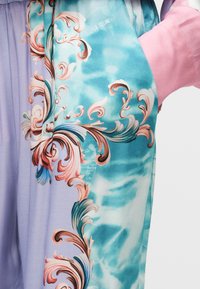 Κοντινό πλάνο υφάσματος με παστέλ μπλε και μωβ μοτίβα ταϊ-νταϊ, διακοσμημένο με περίτεχνα ροζ και τιρκουάζ μπαρόκ ανθικά σχέδια, φορεμένο με ορατό ροζ μανίκι.