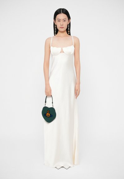 Sieviete baltā, bezroku, līdz grīdai sniedzošā kleitā, turot tumši zaļu sirds formas rokas somiņu ar oranžu un zilu dizainu.