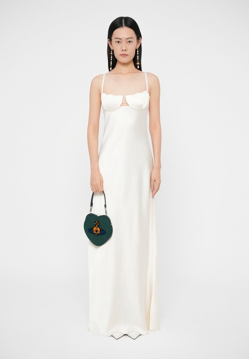 Vrouw in een wit, mouwloos, tot op de vloer reikend jurkje die een donkergroen, hartvormig handtasje vasthoudt met een oranje en blauwe print.