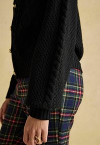 Czarny sweter z dzianiny o splocie warkoczowym na rękawach, zestawiony z spodniami w kratę w kolorach zielonym, niebieskim i czerwonym. Widoczny detal struktury tkaniny.