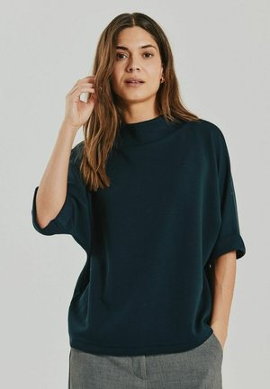 Vrouw met lang bruin haar, gekleed in een donker oversized top en grijze broek, staand met één hand bij haar gezicht en de andere in haar zak.