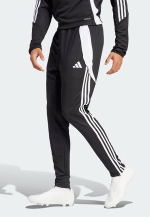 Mand iført sorte Adidas træningsbukser med hvide striber og logo, kombineret med hvide fodboldstøvler, gående mod en lys baggrund.