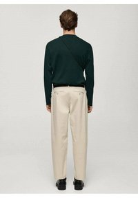 Homme de dos portant une chemise à manches longues vert foncé, un pantalon beige avec ceinture, des chaussures noires et une bandoulière de sac en travers.