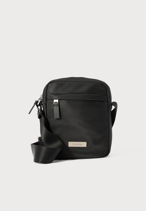 Black nylon crossbody bag with a rectangular shape, adjustable strap, front zip pocket, and silver logo plate on the side.