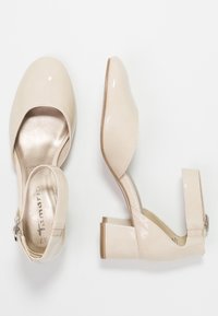 Bežové lakované lodičky s hranatou špičkou, páskem kolem kotníku a blokovým podpatkem. Hladký povrch s lesklým dokončením a tónovými švy.