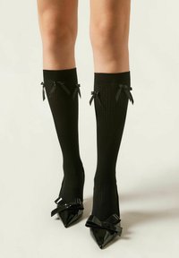 Schwarze gerippte knielange Socken mit zwei kleinen schwarzen Satinbögen am oberen Rand, kombiniert mit spitzen schwarzen Lackabsätzen, die große Schleifen aufweisen.