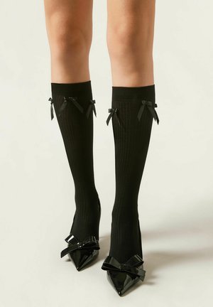 Zwart geribbelde kniehoge sokken met twee kleine zwarte satijnen strikjes aan de bovenkant, gecombineerd met zwarte patent leren pumps met een spitse toe en grote strikken.