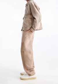 Chaqueta de mezclilla beige con dos bolsillos frontales, llevada sobre pantalones sueltos beige a juego; zapatillas blancas con suelas de un beige claro.