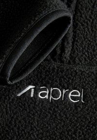 Tissu de veste en polaire noire à texture douce ; comprend une bordure noire et un logo brodé blanc "aprel." Vue en gros plan mettant en valeur les détails de la couture.