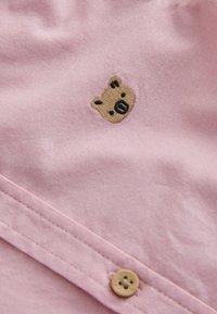 Rožinės medvilnės audinys su išsiuvinėta mažo rudą lokį vaizduojančia veido detalė, šalia šviesiai rudos spalvos sagos su keturių skylių dizainu.