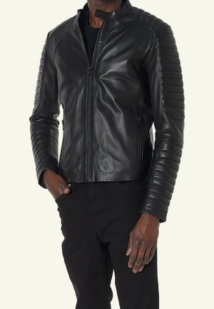 Mężczyzna w czarnej skórzanej kurtce motocyklowej z wyściełanymi rękawami i kieszeniami na suwak, w połączeniu z czarnymi spodniami na jasnym tle.