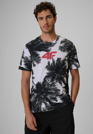 Mężczyzna z kręconymi włosami, ubrany w czarno-biały t-shirt z nadrukiem palm i czerwonym tekstem "Stay Chill" na piersi oraz czarne spodnie.