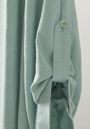 Helles blaus, strukturiertes Stoffhemd mit hochgekrempelten Ärmeln, das über Knopfakzente und ein weiches, lässiges Design verfügt. Sichtbare Nähte.