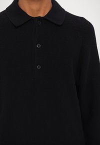 Gros plan d'une personne portant un polo en maille noire avec un col et trois boutons noirs, montrant le cou et le haut du torse.