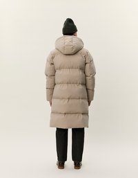 Persona che indossa un lungo cappotto puffa beige con cappuccio, pantaloni neri, scarpe marroni e un berretto verde scuro, in piedi di spalle su uno sfondo neutro.