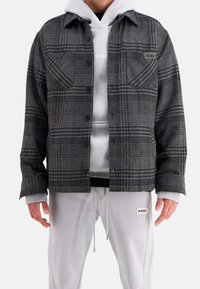 Gråternet flanneljakke med to frontlommer og sorte knapper, lagdelt over en lys grå hættetrøje og parret med lysegrå sweatpants.