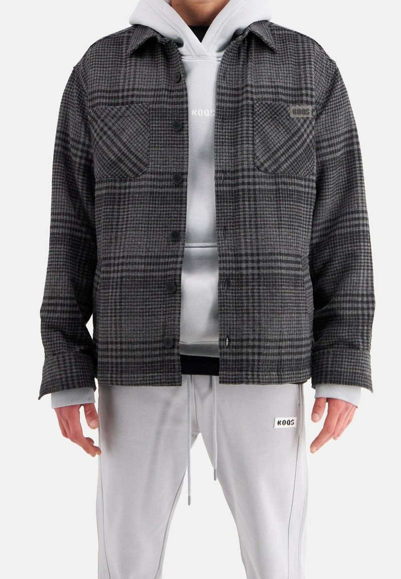 Gråternet flanneljakke med to frontlommer og sorte knapper, lagdelt over en lys grå hættetrøje og parret med lysegrå sweatpants.