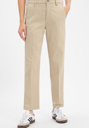 Persona che indossa pantaloni beige a gamba dritta con tasche, abbinati a sneakers Adidas bianche e nere su sfondo uniforme.