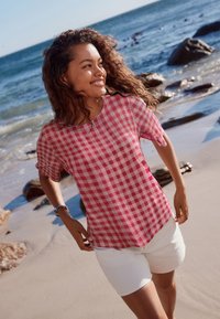 Vaaleanpunavalkoruutuinen lyhythihainen paita pyöreällä kaula-aukolla yhdistettynä valkoisiin shortseihin, rantataustaa vasten.