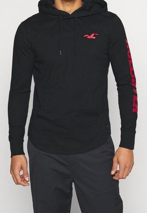 Homme portant un sweat à capuche noir avec un logo de mouette rouge sur la poitrine et le texte « HOLLISTER » sur la manche droite, associé à un pantalon foncé.
