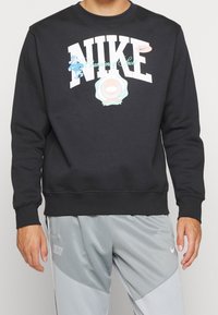 Hombre con sudadera negra de Nike con texto gráfico y logotipos, combinada con pantalones deportivos grises claros de Nike.