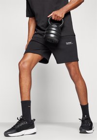 Personne portant une tenue de sport noire tient un kettlebell noir d'une main, debout les jambes écartées sur un sol gris avec un fond blanc.