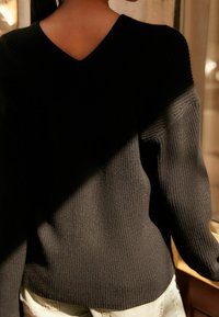Pull noir en maille avec un col en V, présentant une texture côtelée et des manches longues. Le dos arbore un design minimaliste, mettant en avant la simplicité.