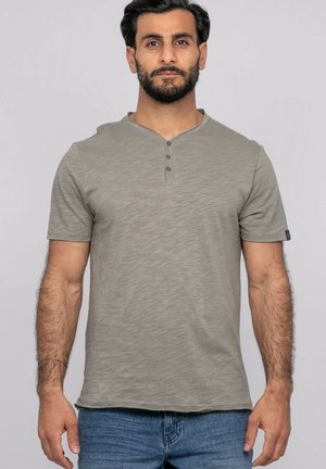 Homme debout face à l'avant portant un t-shirt henley gris à manches courtes et un jean bleu sur un fond clair uni.