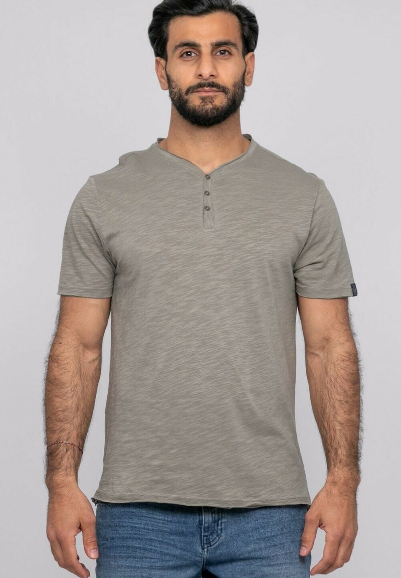 Homme debout face à l'avant portant un t-shirt henley gris à manches courtes et un jean bleu sur un fond clair uni.