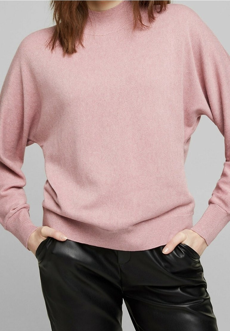 Femme portant un pull en maille rose doux avec des poignets et un col côtelés, associée à un pantalon en cuir noir, les mains dans les poches.