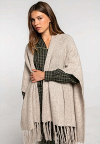 Chal beige de punto con bordes con flecos, tejido texturizado drapeado sobre una camisa a cuadros en verde oscuro y negro. Corte relajado con apertura frontal.