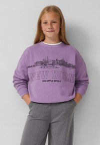 Felpa viola con una grafica dello skyline della città, con "NEW YORK" in caratteri neri e in grassetto e polsini a costine. Indossata con pantaloni grigi.