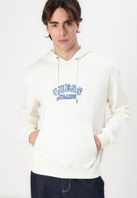 Sudadera blanca con gráfico en azul "GUESS JEANS", cordones, bolsillo frontal, puños y dobladillo de canalé. El modelo lleva puestos jeans de mezclilla oscura.