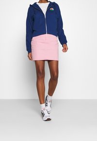 Námořnická zipová mikina s barevným logem, spárovaná se světle růžovou džínovou sukní. Model má na sobě bílé tenisky a ponožky.