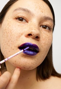 Colore labbra viola intenso con finitura lucida applicato con un pennello. La texture della pelle con lentiggini è visibile; lo sfondo è bianco semplice.