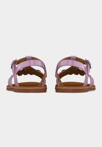 Ζευγάρι επίπεδων καφέ σανδαλιών με λαμπερές βιολετί λουριά και ασημένιες αγκράφες, παρουσιασμένα από πίσω σε λευκό φόντο.