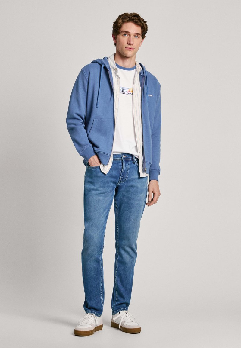 Sudadera con capucha azul, camiseta blanca con logo, camisa de cuadros claros, jeans azul claro, zapatillas blancas con suelas marrones. Conjunto informal, corte holgado.