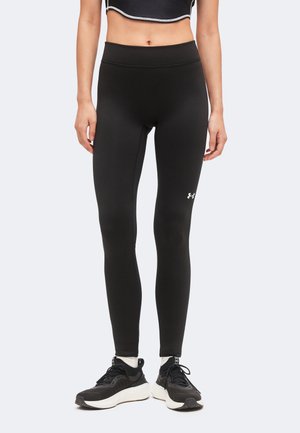 Žena nosiaca čierne legíny Under Armour, čierno-biele tenisky a čierne krátke tričko s bielym lemom, stojaca na jednofarebnom pozadí.