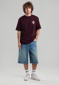 Maroon cotton t-shirt with short sleeves and white logo, paired with light blue denim shorts, white sneakers, and green-striped socks.