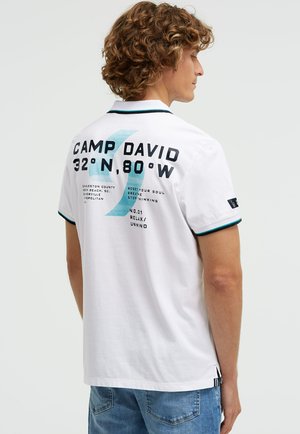 Bărbat tânăr cu păr creț poartă un tricou polo alb cu textul „Camp David 32°N, 80°W” și un grafic albastru pe spate, asortat cu blugi albastre.