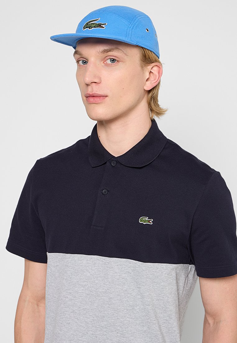 Navy en grijs kleurblock polo shirt met een platte kraag, voorzien van een kleine groene geborduurde krokodil logo op de borst. Lichtblauwe baseballpet.